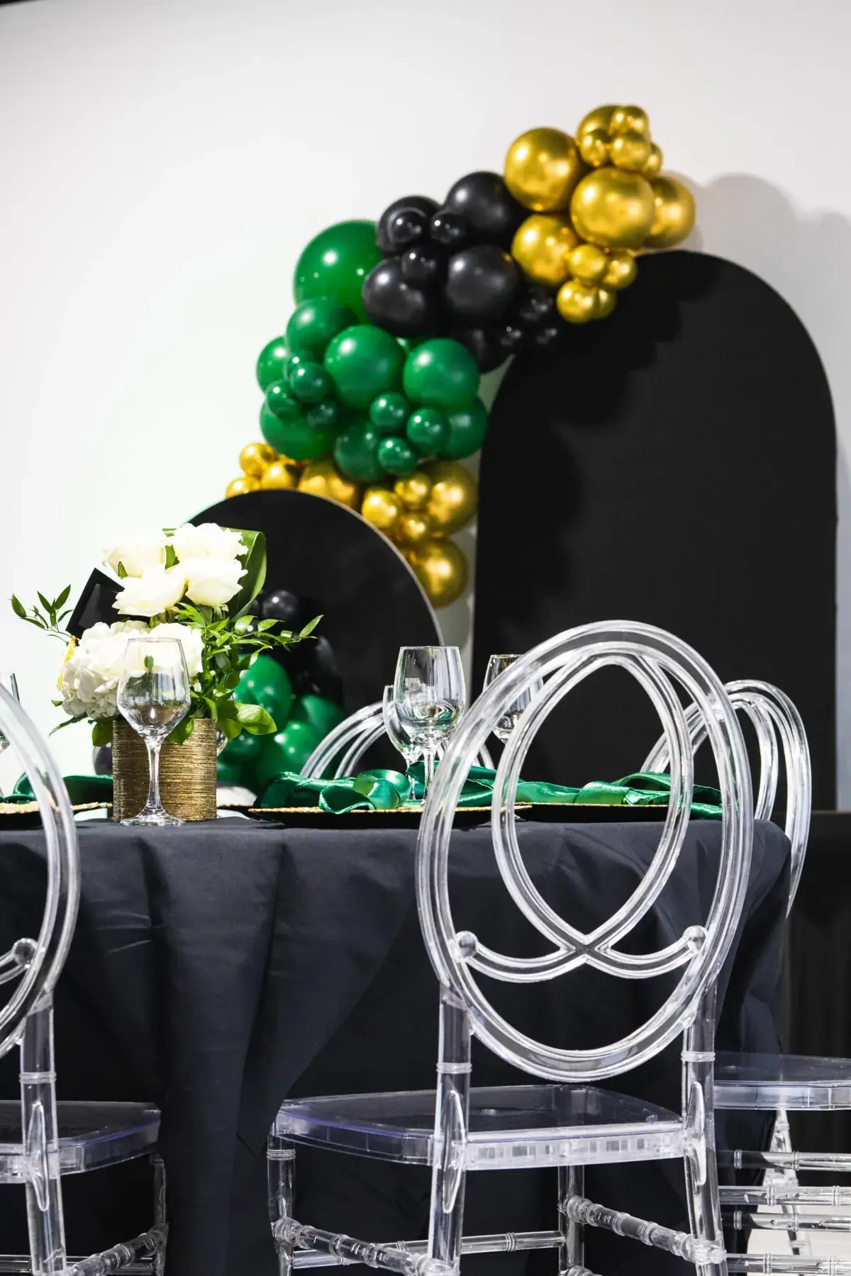Black and gold balloon backdrop with acrylic ghost chairs and black linens set up inside Event Center Miami in Kendall