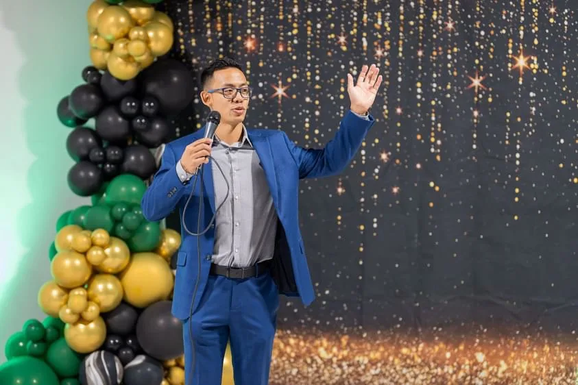 A speaker addressing guests in front of a gold and black balloon garland at Event Center Miami